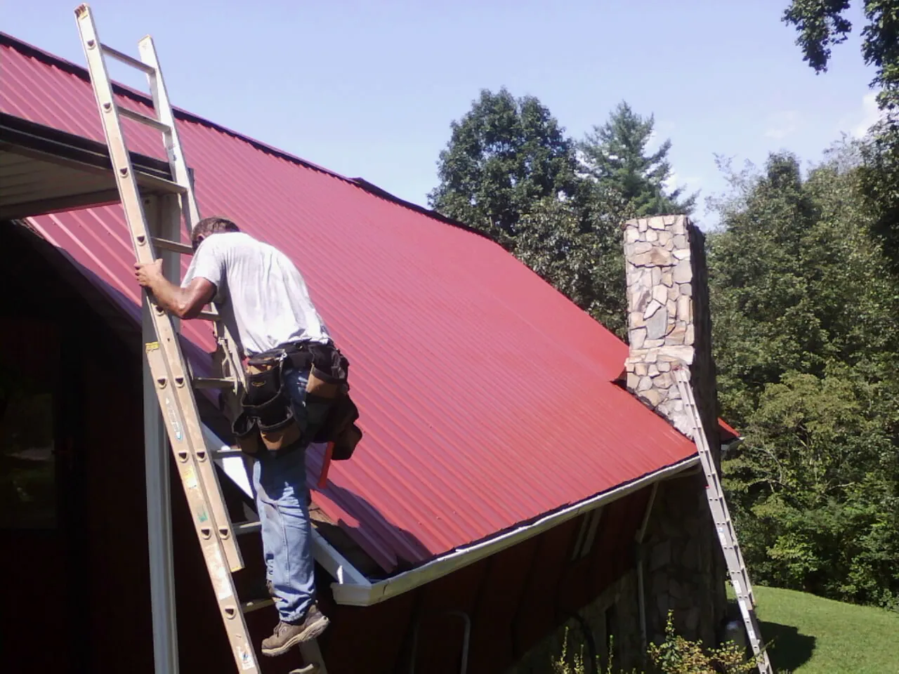 Roofer on a ladder beside a red metal roof and stone chimney — Soffit & Fascia Repair by Roofing Brothers in San Carlos